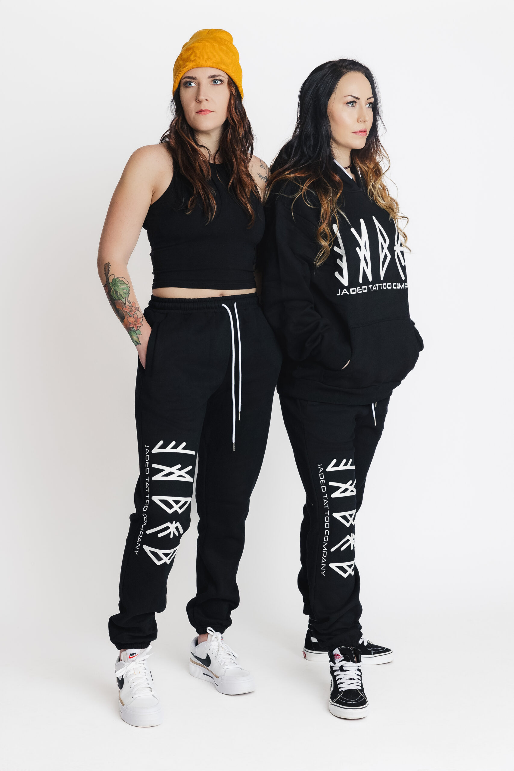 Two women in photo studio modeling merch for Jaded Tattoo Company. Black sweatpants and black hoodie embossed with the Jaded logo